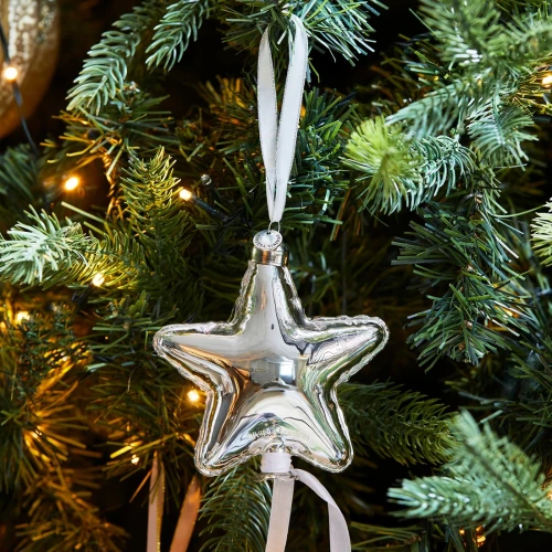 Bombka Balloon Star S Riviera Maison, srebrna gwiazda z metalicznego materiału, zawieszona na białej tasiemce, umieszczona na zielonym drzewku wśród światełek z widocznym logo na dole.