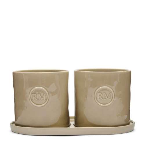 Osłonki na zioła w zestawie Portofino Herbs Set Riviera Maison, w kolorze jasno beżowym, ceramiczne, z wyraźnym logo „RM” na przedniej stronie. Dwa stojące na podstawce pojemniki o zaokrąglonych krawędziach, gładkiej powierzchni.