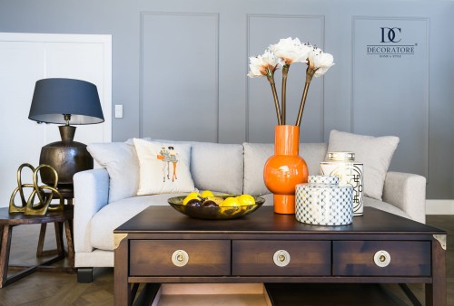 Szara, rozłożona sofa trzyosobowa w tkaninie Gemma z poduszką dekoracyjną. Na brązowym stoliku czarna misa z owocami, pomarańczowy wazon z kwiatami i ceramiczne pojemniki, na tle w odcieniu szarym.