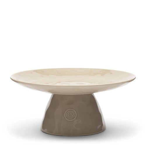 Beżowa patera na ciasto Riviera Maison Portofino z ceramicznym, połyskującym talerzem na okrągłej podstawie z logo RM. Stojąca na niskiej, stabilnej nodze, idealna do serwowania ciast i deserów.