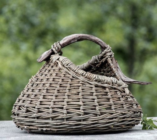 Podręczny koszyk Rustic Rattan o zaokrąglonym kształcie, wykonany z plecionego rattanu w odcieniach szaro-brązowych, z uchwytem z naturalnego drewna. Koszyk leży na stole, w tle delikatne, zielone tło.