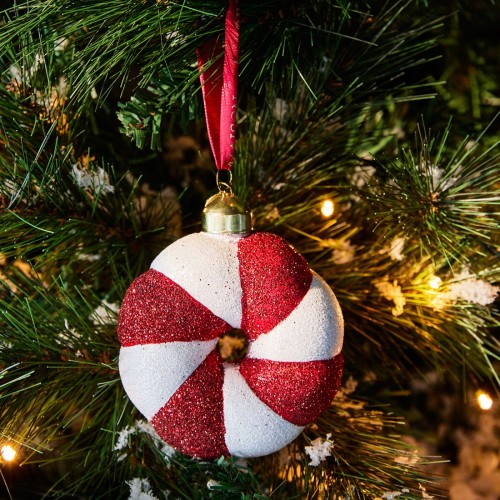 Zawieszka choinkowa o średnicy 10 cm w kształcie cukierka z czerwonymi i białymi pasami, pokryta brokatem. Złoty uchwyt i czerwona wstążka do zawieszenia. Umieszczona na gałęzi choinki, w tle widoczne delikatne światełka.