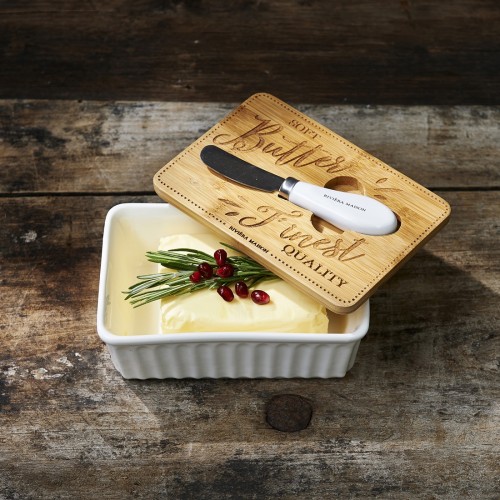 Maselniczka z białej ceramiki z pokrywą z drewna bambusowego, na której widnieje napis Finest Quality Butter. W środku znajduje się kostka masła ozdobiona gałązkami rozmarynu i czerwonymi owocami. Obok leży nóż do masła.