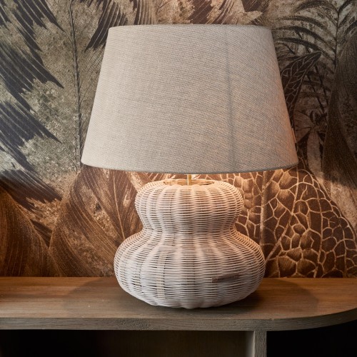 Rattanowa podstawa lampy Valenton w kolorze białym, o kształcie przypominającym kulę, z jasnoszarym abażurem w kształcie stożka. Lampa ma wysokość 43 cm, stoi na drewnianym blacie, w tle widoczny wzór liści.