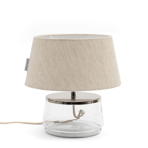 Lampa stołowa Piola Fillable Riviera Maison o wysokości 39 cm. Przezroczysta szklana podstawa z metalowym elementem i naturalnym, jasnobeżowym kloszem z tkaniny. Kabel z wtyczką widoczny z boku.