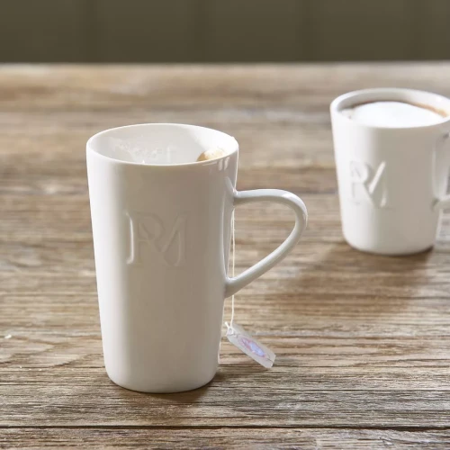 Biały kubek Monogram Riviera Maison XL z lekko zaokrąglonym brzegiem i wyprofilowaną rączką. Na powierzchni widoczny subtelny wzór RM. Produkt ustawiony na drewnianym stole, z widoczną torebką herbaty.