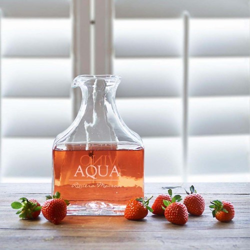 Karafka Riviera Maison Vino Aqua w szklanym, prostokątnym kształcie z uchwytem, napełniona różowym płynem. Na przedzie czytelny napis Vino Aqua. Produkt umieszczony na drewnianym blacie, obok świeże truskawki. Jasne, rozmyte tło z oknami.