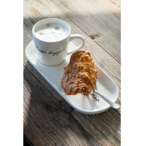 Biały kubek z napisem Caffe Doppio zawiera kawę z pianką, umieszczony na prostokątnej talerzyku. Obok znajduje się mały, chrupiący wypiek z migdałami. Tło to drewniany stół, co podkreśla przytulny klimat.