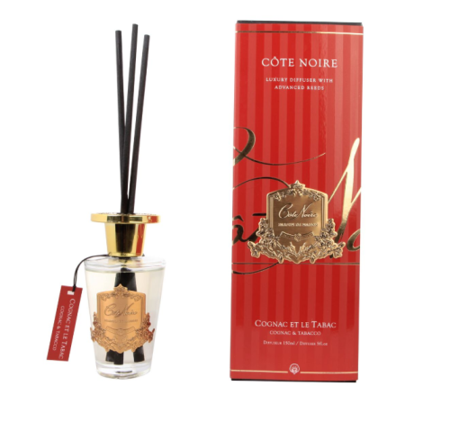 Dyfuzor zapachowy Cote Noire Cognac&Tabacco w eleganckiej butelce z przezroczystego szkła, z złotą zakrętką i trzema czarnymi patyczkami. Opakowanie w czerwono-złotej kolorystyce z logo Côte Noire. Pojemność 150 ml, widoczna etykieta z nazwą zapachu.