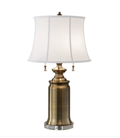 Lampa stołowa Wilington w kolorze mosiądzu z białym abażurem, stojąca, o wysokości 69 cm. Abażur w kształcie stożka, z delikatnie widocznymi pionowymi przeszyciami. Widoczne dekoracyjne detale na podstawie. Minimalistyczne tło.