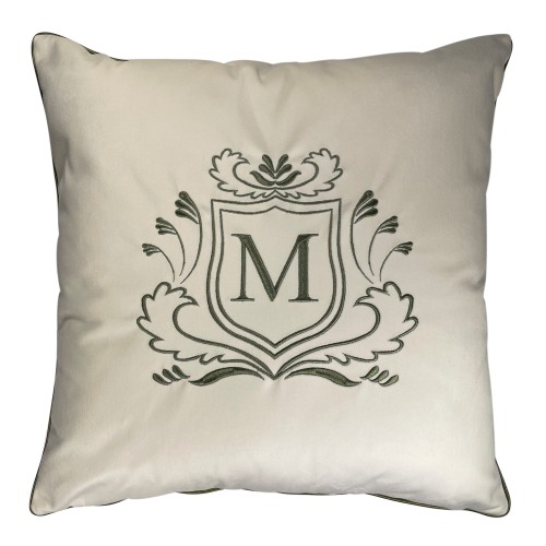 Poduszka haftowana dekoracyjna w kolorze kremowym, wykonana z welwetu. Frontalnie umieszczony monogram M w zielonym owalnym tle otoczonym ornamentalnym haftem. Produkt leży na gładkim tle, ukazując fakturę materiału oraz detale haftu.