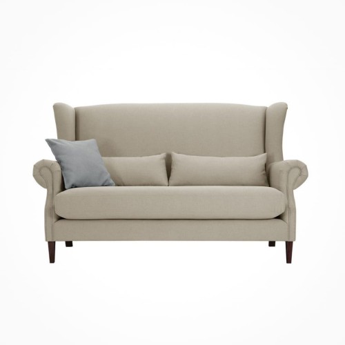 2,5 osobowa sofa klasyczna w jasnobeżowym kolorze z drewnianymi nóżkami. Ma wysokie oparcie z charakterystycznymi bocznymi podłokietnikami, poduszki oparciowe w zestawie. Ujęcie frontalne, stojąca w na jednolitym białym tle.