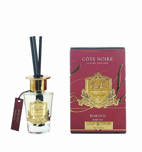Dyfuzor zapachowy Cote Noire Rose Oud w przezroczystym szklanym pojemniku ze złotą pokrywką i trzema czarnymi patykami. Obok widoczny kartonowy pudelko w burgundowym kolorze z złotymi detalami i napisem 'Rose Oud'. Produkt stoi na jasnym tle.