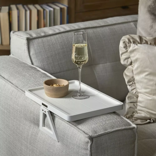 Tacka Monogram Plateau De Fauteuil Riviera Maison w kolorze białym, umieszczona na oparciu szarej sofy. Na tacce znajduje się szklanka z szampanem i drewniana miseczka z orzeszkami. Ujęcie z boku, pokazujące detal montażu.