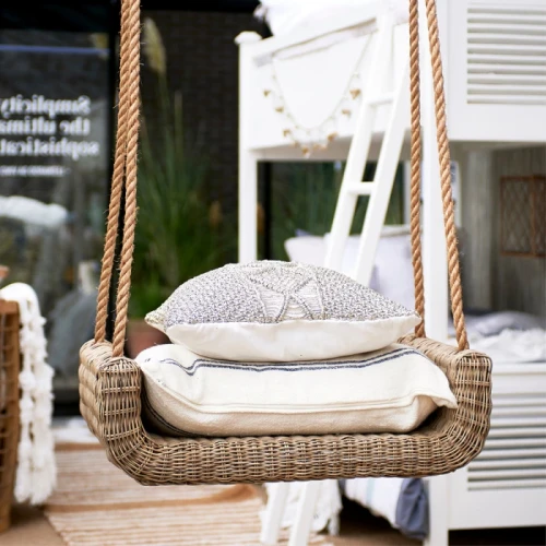 Rattanowa huśtawka w naturalnym kolorze na linach, z dwoma poduszkami: dekoracyjną i jasną. W tle stylowa aranżacja z białymi meblami i roślinami podkreślająca design.
