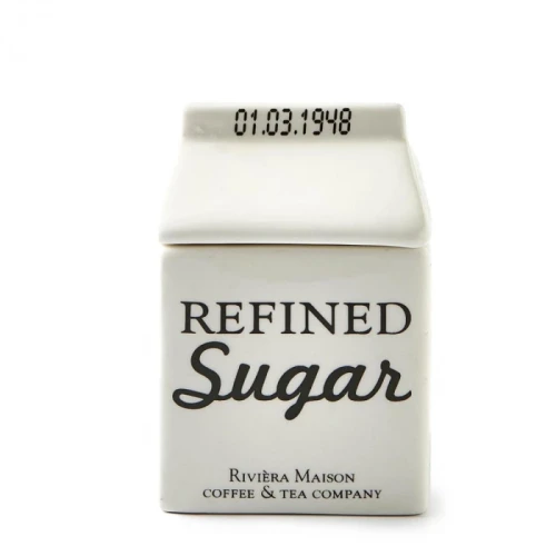 Biała cukierniczka o kształcie małej kartonowej paczki, z napisem REFINED Sugar w czarnym kolorze. Produkt prezentowany frontalnie, z otwartym dnem. Widoczne logo Riviera Maison na dolnej części. Gładka, szkliwiona powierzchnia.