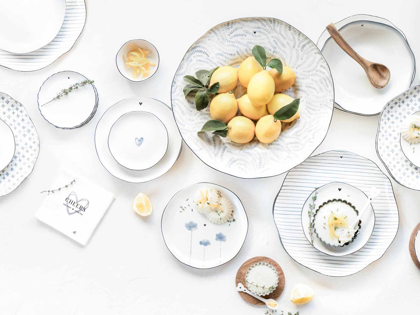 Talerz śniadaniowy Iris Blue Print z kolekcji Bastion Collection w kolorze błękitnym, z delikatnym wzorem i ciemnoniebieską linią na brzegu. Produkt leży na białym tle obok innych talerzy, z dodatkami cytryn i ziół. Widoczna faktura porcelany.