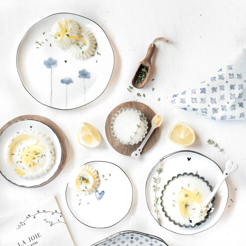 Talerz głęboki Iris Blue Bastion Collection w biało-niebieskiej kolorystyce z delikatnym kwiatowym wzorem. Produkt leży na białym tle, obok znajdują się cytryny, zioła, oraz łyżeczki. Talerz ma falowane brzegi i wyraźny detal w postaci serca.