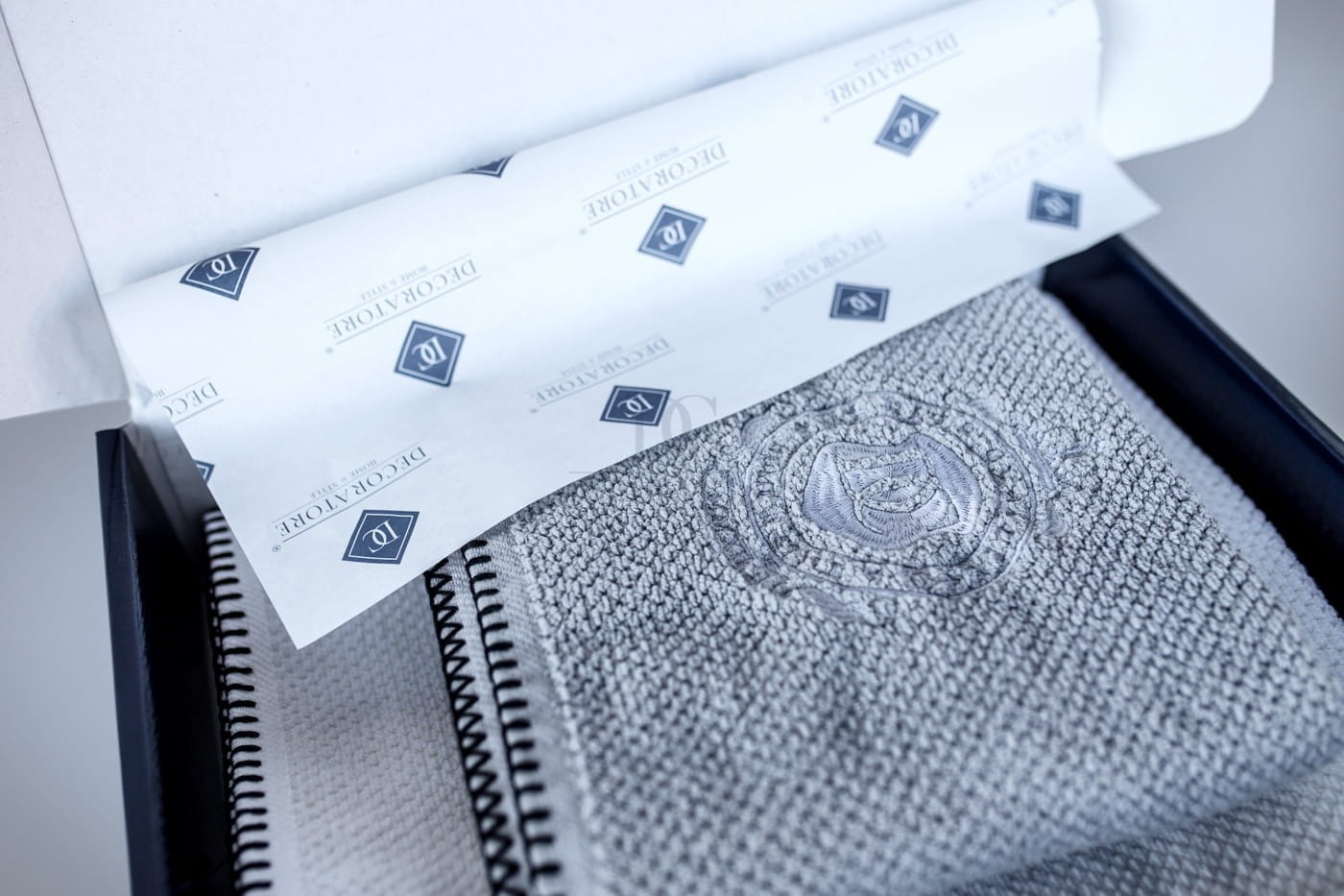 Personalizowany ręcznik w białym kolorze, o ryżowej strukturze, z haftem, prezentowany w otwartej, eleganckiej opakowaniu. Widoczna faktura materiału oraz logo na papierze. Produkt leży wewnątrz pudełka, ukazując detale haftu.