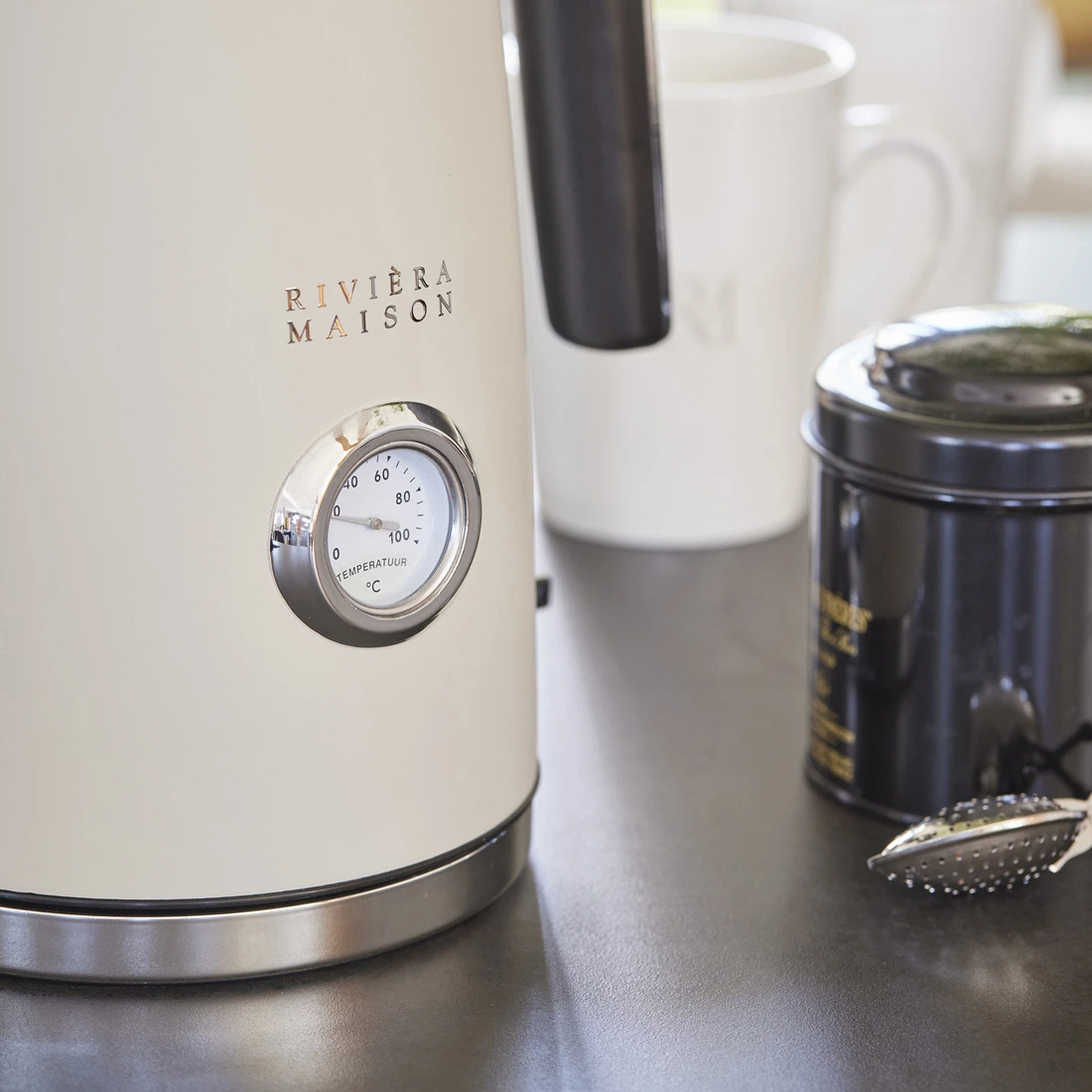 Czajnik Classic Riviera Maison w kolorze kremowym, z metalowym wykończeniem. Z boku znajduje się analogowy wskaźnik temperatury do 100°C. Produkt umieszczony na ciemnym blacie, w tle widoczne dwa kubki oraz czarna puszka z herbatą.