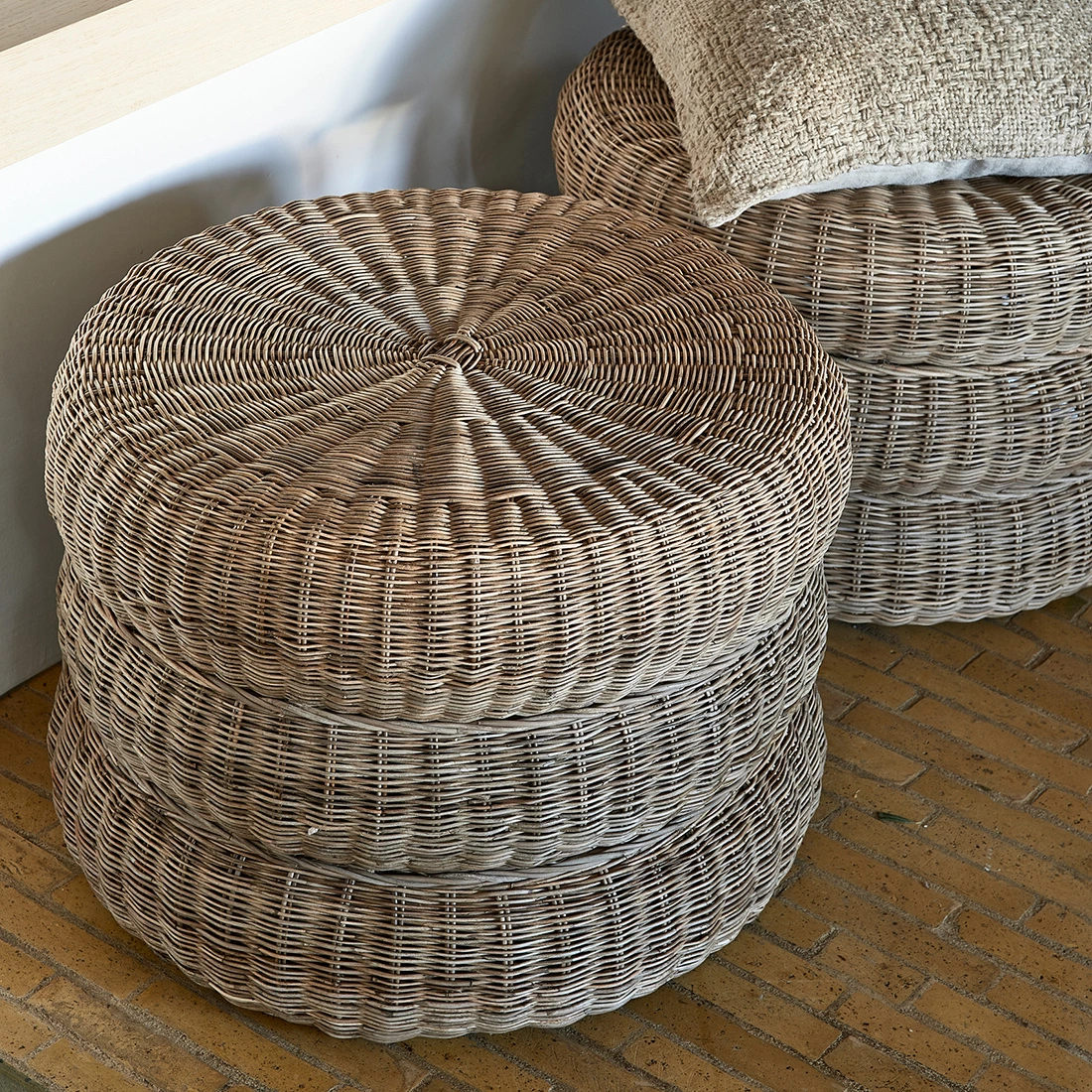 Puf Rustic Rattan w kolorze jasnego beżu, o okrągłym kształcie, wykonany z plecionki rattanowej. Produkt ułożony w stos na jasnej podłodze, z widoczną teksturą materiału. Na górze znajduje się jasna poduszka dekoracyjna.