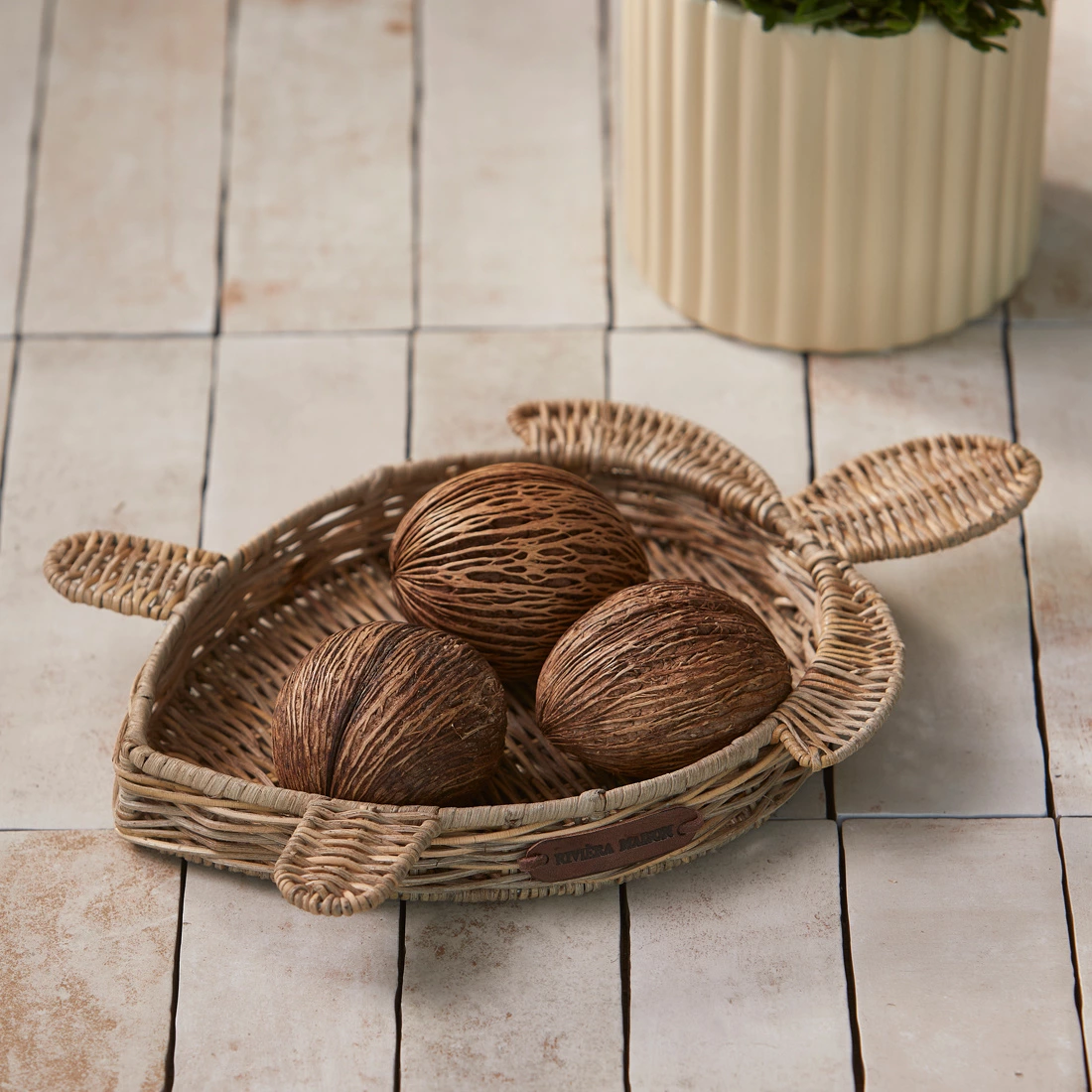 Rattanowa tacka do serwowania w kształcie żółwia, w kolorze naturalnym, z dwoma uchwytami. Widoczne brązowe dekoracyjne kule. Produkt leży na jasnym tle, w aranżacji z doniczką w tle. Wymiary i detale struktury rattanowej niewidoczne.