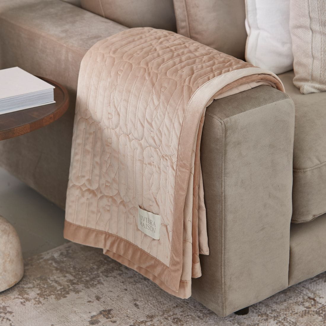 Stylowy pled Loreo 180x130 cm w kolorze delikatnego różu, z teksturą pikowaną. Produkt leży na oparciu szarej sofy. W rogu widoczna metka z logo Riviera Maison. Tło w neutralnych odcieniach.