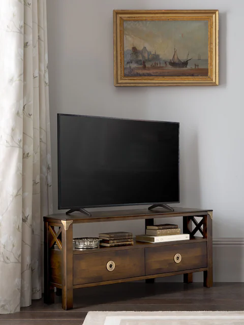 Stolik narożny RTV drewniany w kolorze kasztanowym, o wymiarach 110x40x50 cm, z dwiema szufladami i ozdobnymi elementami na bokach. Produkt widoczny z boku, z telewizorem na blacie, umieszczony w stylowym wnętrzu.