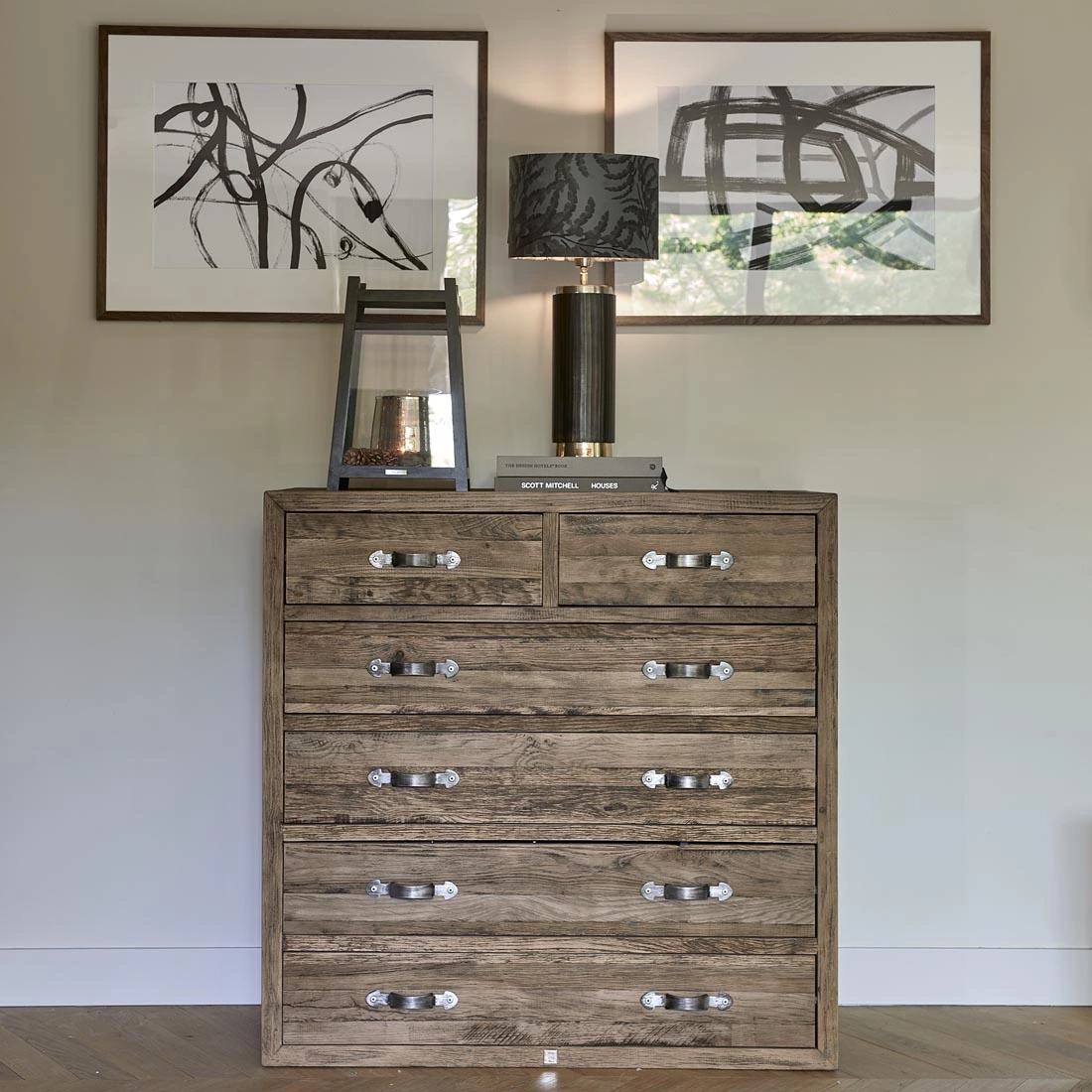 Komoda Connaught XL w kolorze dębu, z sześcioma szufladami z metalowymi uchwytami. Ujęcie frontalne, produkt stoi w jasnym wnętrzu, obok dekoracyjnych lamp i obrazów. Powierzchnia matowa, prostokątny kształt, wymiary optymalne do przechowywania.
