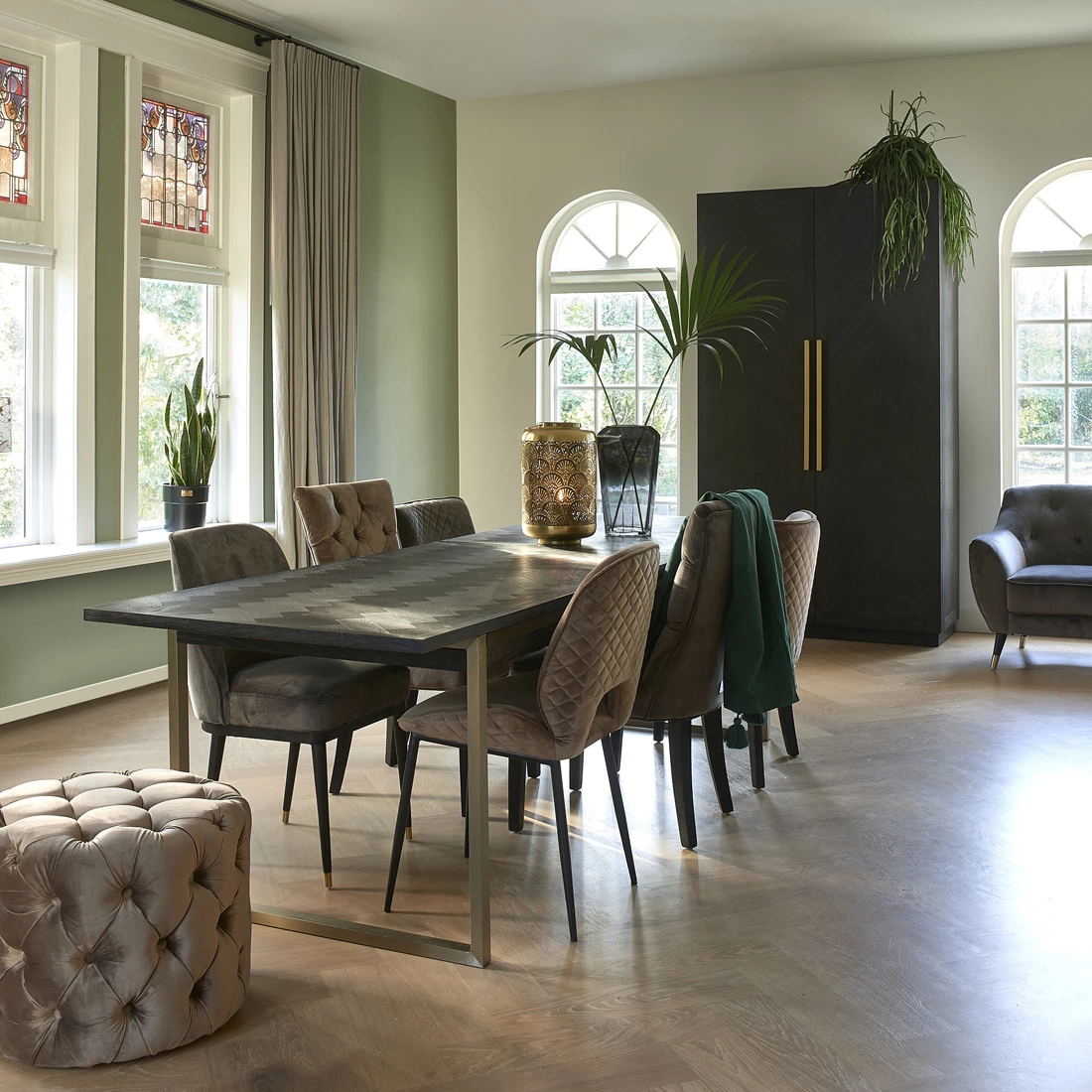 Kredens 5th Avenue Riviera Maison w ciemnym kolorze o prostych liniach i złotych uchwytach. Produkt stoi w jasnym wnętrzu, obok drewnianego stołu i tapicerowanych krzeseł. W tle okna z witrażami oraz rośliny doniczkowe.