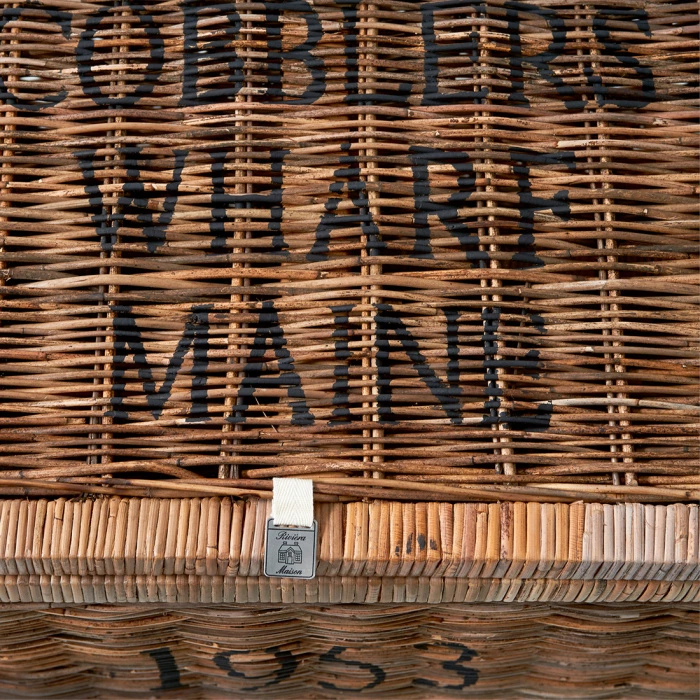 Rattanowy kufer w jasnym kolorze z widocznym napisem COBBLERS WHARF MAINE o intensywnej czerni, z metalowym zamknięciem. Ujęcie z góry podkreśla jego teksturę i detal konstrukcji. Produkt w nowym stanie, przeznaczony do przechowywania.