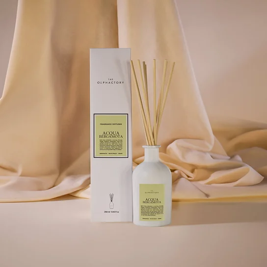 Dyfuzor zapachowy Insignia Acqua Bergamota 250 ml w białej, zaokrąglonej butelce z naturalnymi patyczkami umieszczony obok eleganckiego opakowania. Tło w neutralnym kolorze, lekko teksturowanym, podkreśla minimalistyczny wygląd produktu.