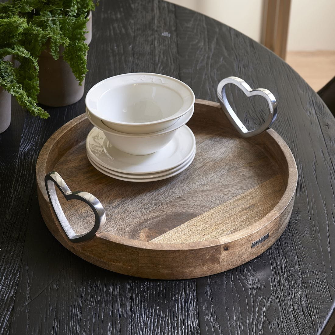 Taca Pretty Hearts Riviera Maison, okrągła, w kolorze naturalnego drewna, z metalowymi uchwytami w kształcie serca. Na tacy znajdują się białe talerze oraz miski. Produkt leży poziomo na czarnym stole, w otoczeniu roślinności.