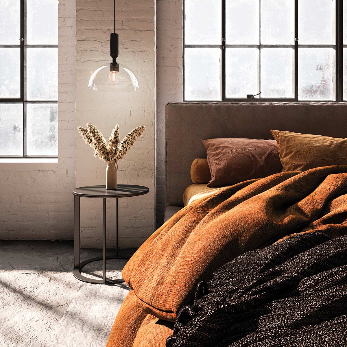 Czarna lampa wisząca Jude z przezroczystym kloszem, zamontowana nad niskim stolikiem w sypialni. Produkt wiszący z kablem w kolorze black. Obok stylowe dekoracje i poduszki w odcieniach brązu i beżu, na tle z jasnej cegły.