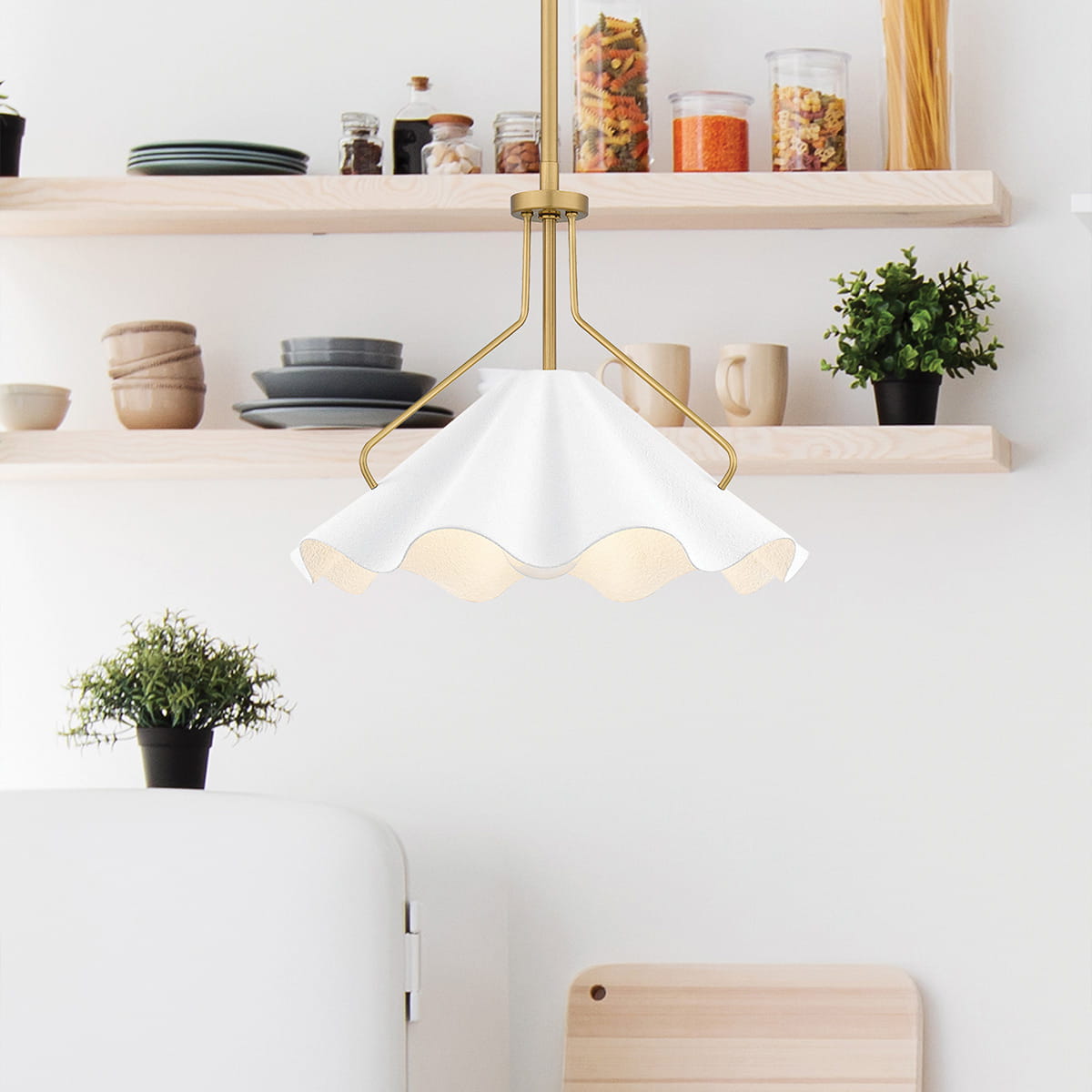 Lampa wisząca Mejia w kształcie falistego klosza, kolor biały z mosiężnymi detalami. Zawieszona na złotym stelażu. Widok frontalny, w kuchennej aranżacji, z półkami w tle, na których znajdują się naczynia i dekoracje.