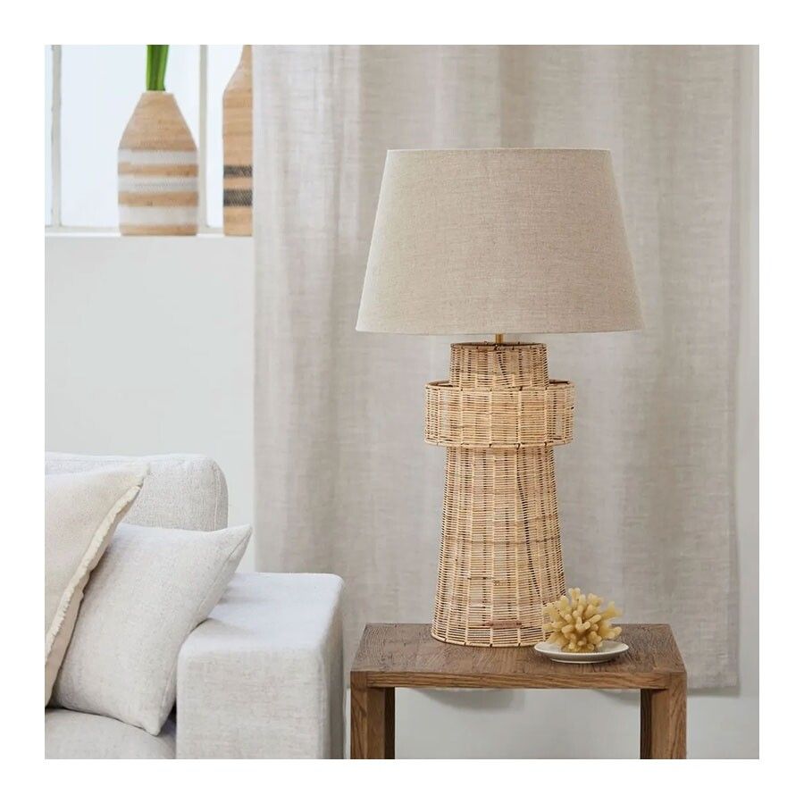 Rattanowa podstawa lampy o wysokości 70 cm w naturalnym kolorze, z okrągłym abażurem z tkaniny. Produkt prezentowany na stojaku w drewnianym stylu, w otoczeniu jasnych zasłon i dekoracyjnych elementów. Stojący, z widoczną teksturą rattanu.