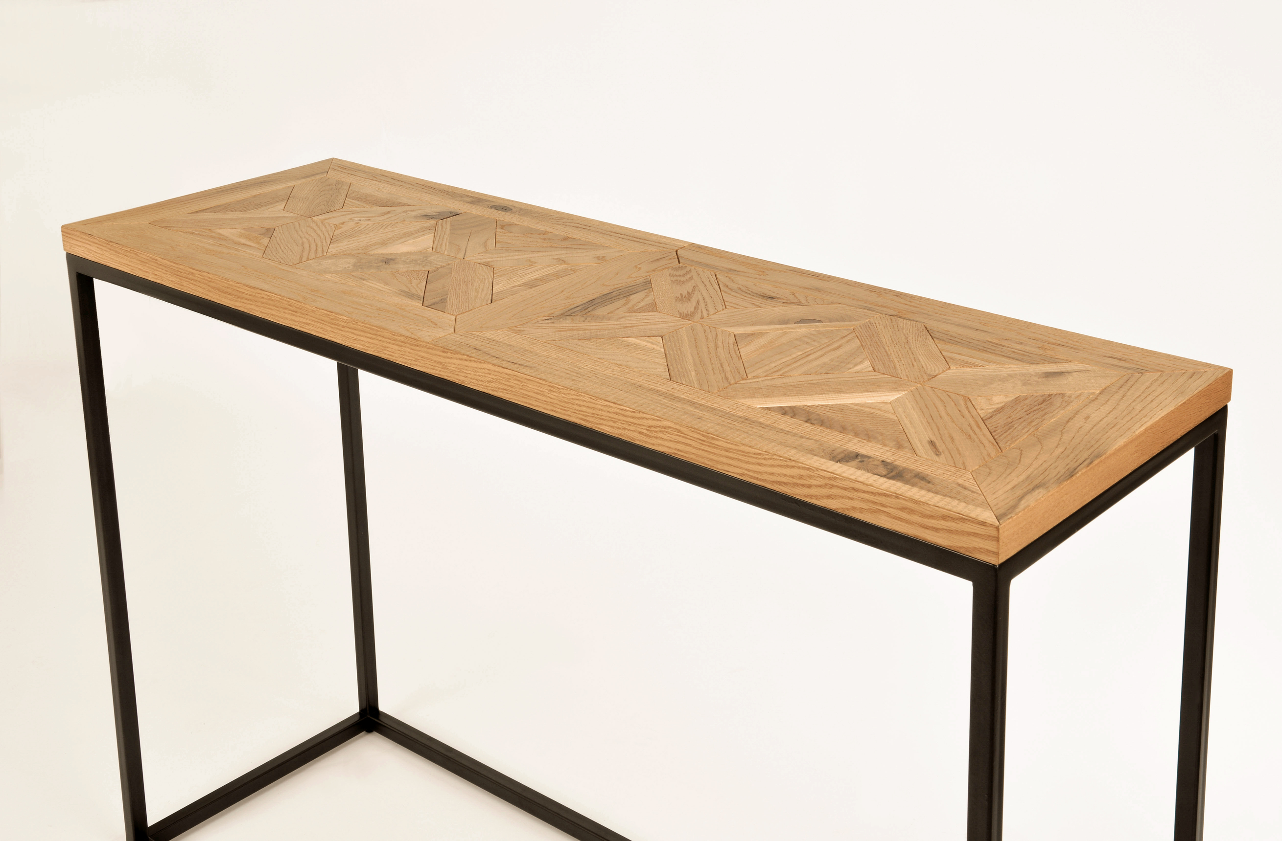 Industrialna konsola o wymiarach 116 cm z naturalnego dębu, w kolorze jasnego drewna z geometrycznym wzorem. Stalowa, matowa rama w ciemnym graficie. Produkt stoi na równym podłożu, widoczny z boku. Prosta, jasna ekspozycja na jednolitym tle.