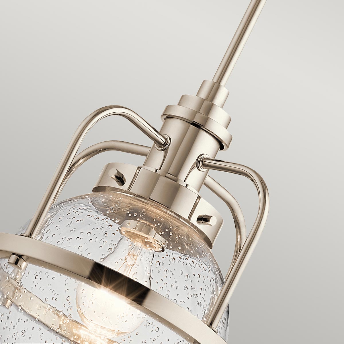 Lampa wisząca Triocent w kolorze polerowanego niklu. Ujęcie z góry pokazuje przezroczystą, kulistą szklankę z widocznymi bąbelkami, metalowe mocowanie oraz dekoracyjne uchwyty. Elementy wzornictwa łączą nowoczesność z elegancją.