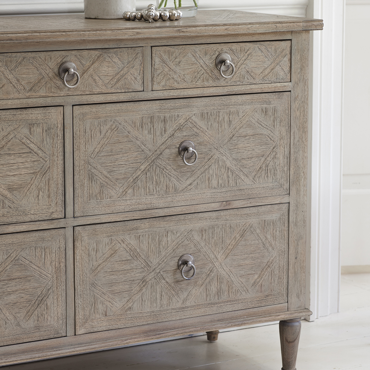 Drewniana komoda Vintage Rustique o wymiarach 130x45x88 cm, w jasnym odcieniu z widocznymi geometrycznymi wzorami na frontach szuflad. Posiada trzy szuflady z okrągłymi uchwytami w kolorze metalicznym. Stojąca na lekko zaokrąglonych nogach.