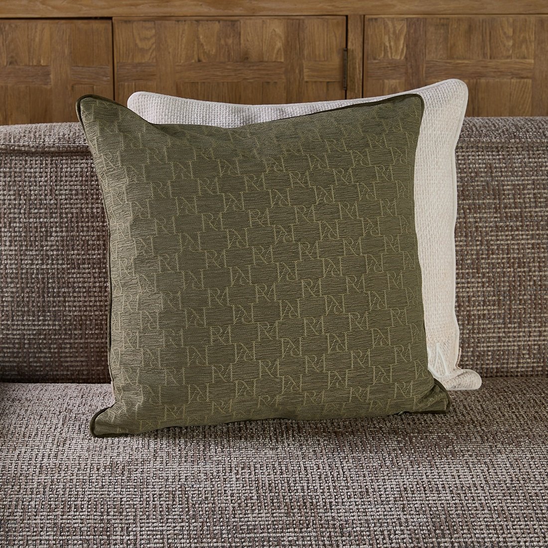 Poszewka RM Monogram Belliere 50x50 w kolorze oliwkowym, z delikatnym wzorem logo RM. Produkt umieszczony na szarej kanapie, z lekko zaokrąglonymi rogami i kontrastową lamówką. W tle widoczna jasna poszewka.