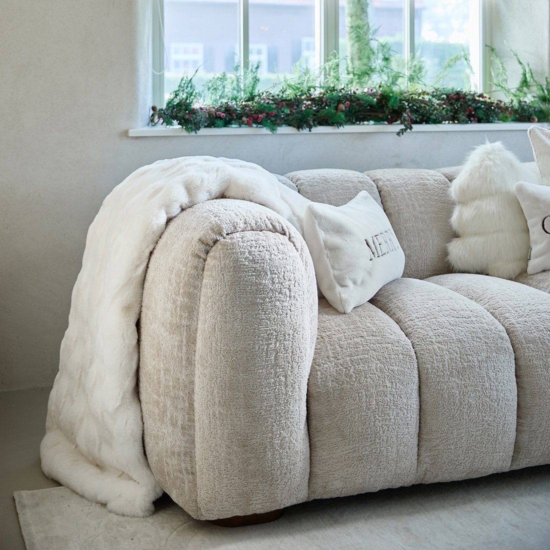 Pled Romolo Faux Fur w kolorze jasnego beżu, o wymiarach 180x130 cm, leży na wygodnej kanapie. Produkt ma miękką, teksturowaną powierzchnię i delikatny połysk. W tle widoczne są poduszki oraz okno z roślinnością na parapecie.