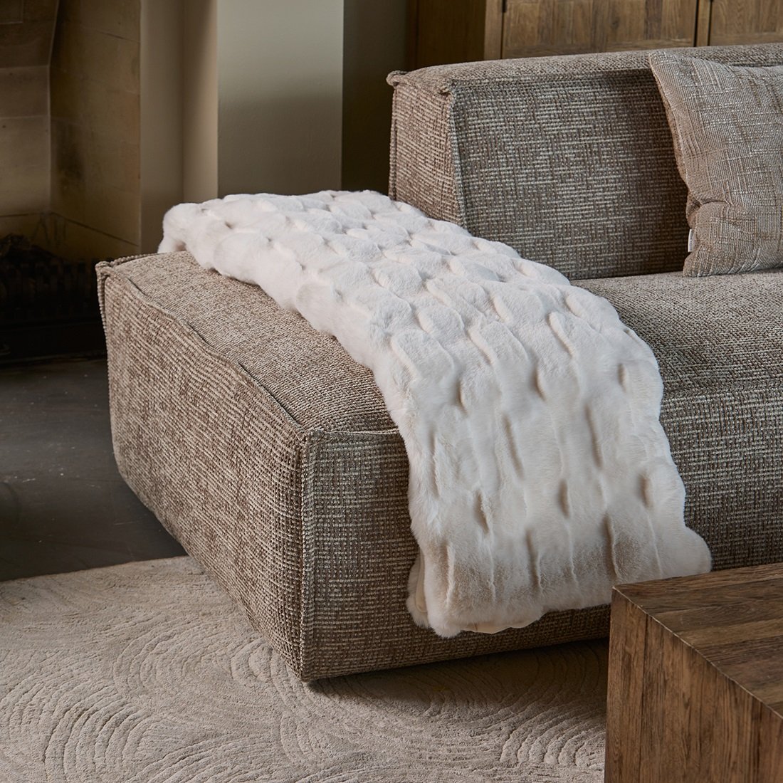 Pled Romolo Faux Fur Riviera Maison w kolorze jasnym, o fakturze imitującej futro, leży na sofie w odcieniach brązu. Produkt ma wymiary 180x130 cm, prezentując delikatny wzór i miękkość, idealny do dekoracji wnętrz.