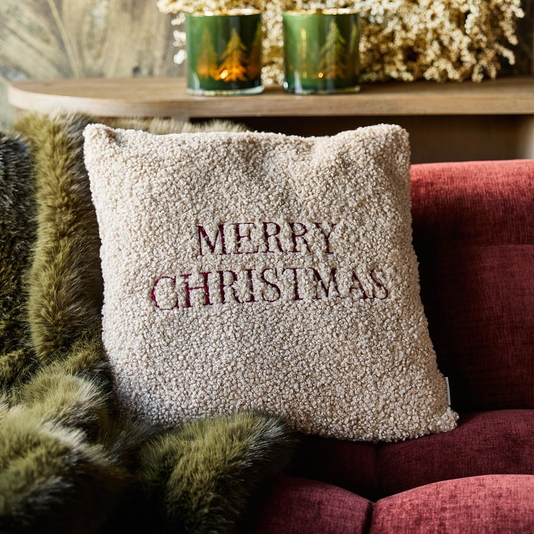 Poszewka 50x50 cm w kolorze kremowym z haftem MERRY CHRISTMAS w bordowym odcieniu. Produkt leży na czerwonym tle, obok widoczna zielona narzuta i dekoracyjne świeczniki. Tekstura materiału jest miękka i puszysta.