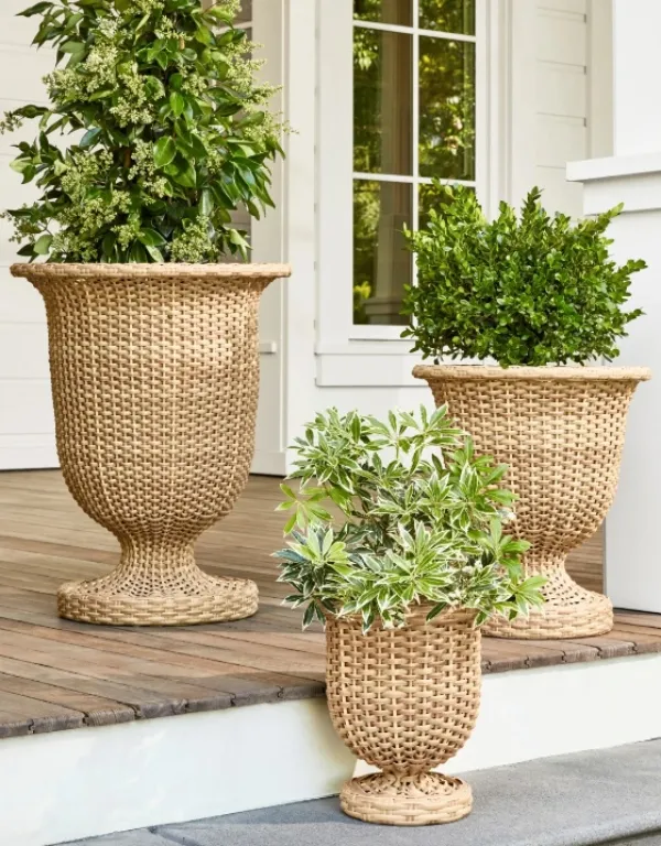 Wazon rattanowy Charlotte o wysokości 50 cm, w naturalnym kolorze, z plecionką w stylu vintage. Stojący na tarasie wśród roślin, z okrągłą podstawą i ozdobnym brzegiem. Idealny do dekoracji przestrzeni zewnętrznych.