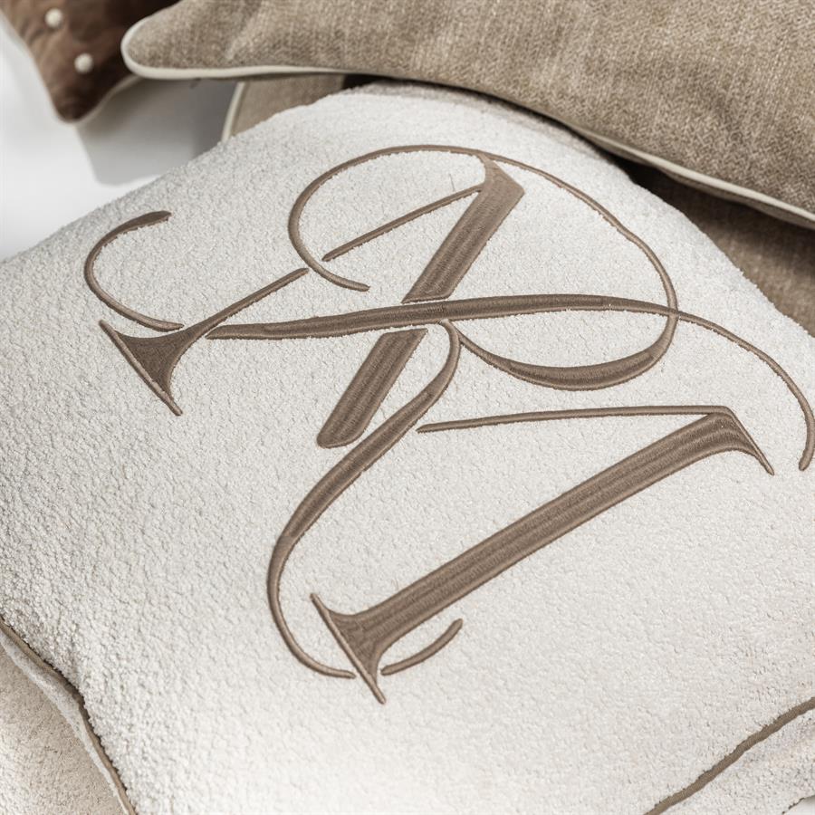 Poduszka dekoracyjna Bouclé w kolorze białym o wymiarach 60x60 cm z haftowanym logo w odcieniu brązowym. Tekstura materiału jest miękka i puszysta. Produkt leży płasko na tle innych poduszek.