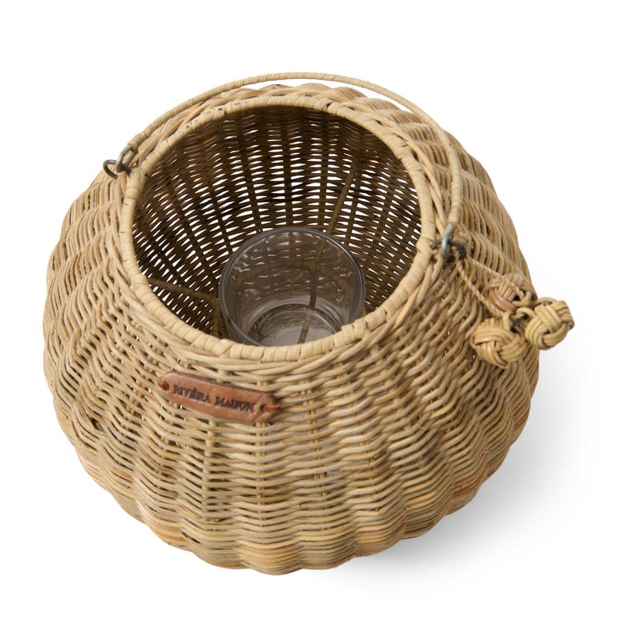 Rattanowy lampion Cherchio w kolorze naturalnym, o zaokrąglonym kształcie, z widocznym szklanym wkładem wewnętrznym. W górnej części uchwyt do zawieszenia oraz drewniana etykieta z logo Riviera Maison. Produkt leżący na białym tle.
