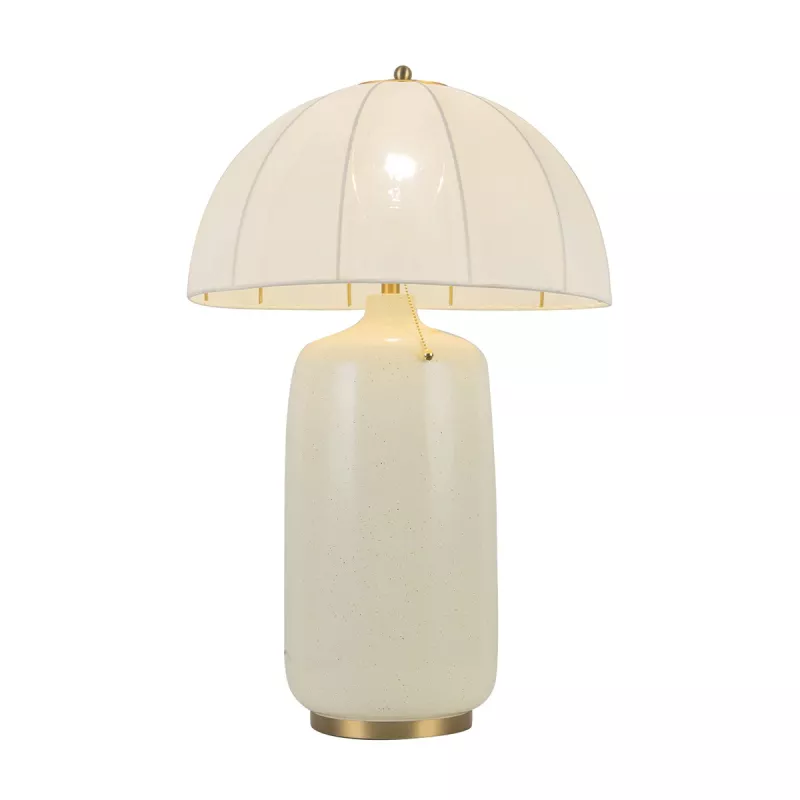 Lampa stołowa Crosby w szczotkowanym złocie z okrągłym, białym kloszem. Produkt ma ceramiczną, matową podstawę w jasnym kolorze oraz złotą, metalową podstawę. Ujęcie frontalne, lampa stoi na płaskiej powierzchni.