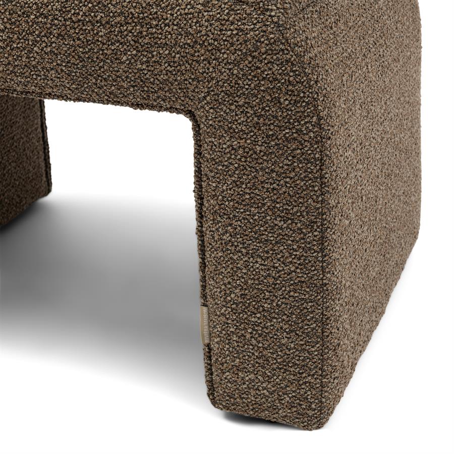 Puf Brera Boucle Classic Brown Riviera Maison w kolorze brązowym, o prostokątnym kształcie i teksturowanej fakturze materiału. Widoczny detal to zaokrąglony róg oraz subtelna metka na boku. Produkt leży na jasnym tle, w pozycji poziomej.