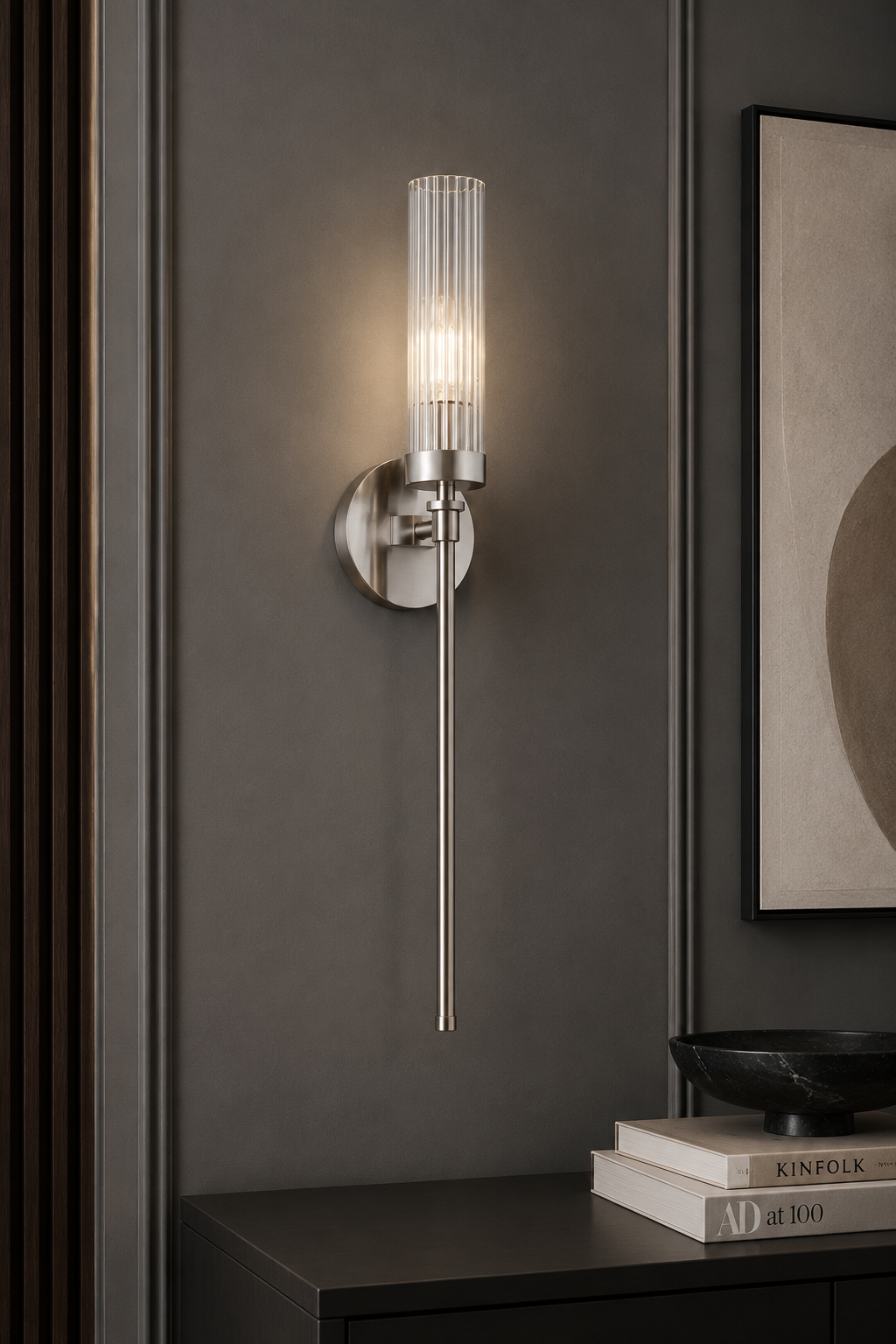 Kinkiet Preston w kolorze niklowym, wysokość 58 cm, z prostokątnym kloszem z przezroczystego szkła, montowany na ścianie. Produkt o eleganckim designie, umieszczony obok stolika z książkami i dekoracyjną misą. Tło w odcieniach szarości.