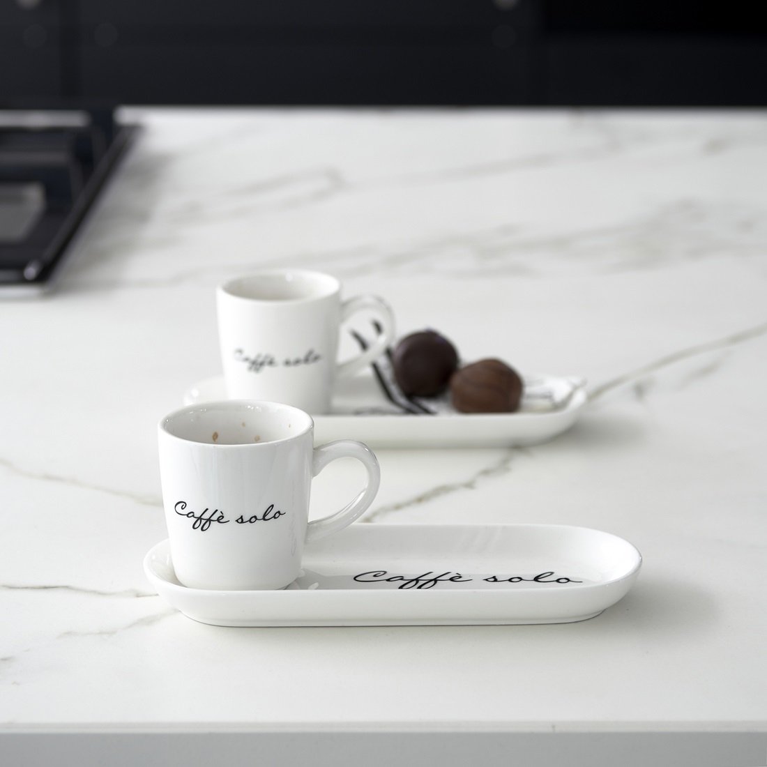 Kubek z podstawkiem w kolorze białym, z napisem Caffè solo w czarnym kolorze. Kubek ma mały uchwyt, a na podstawce znajdują się dwa ciemne kulki. Ujęcie pokazuje produkt na marmurowym blacie, w tle widoczny inny kubek.